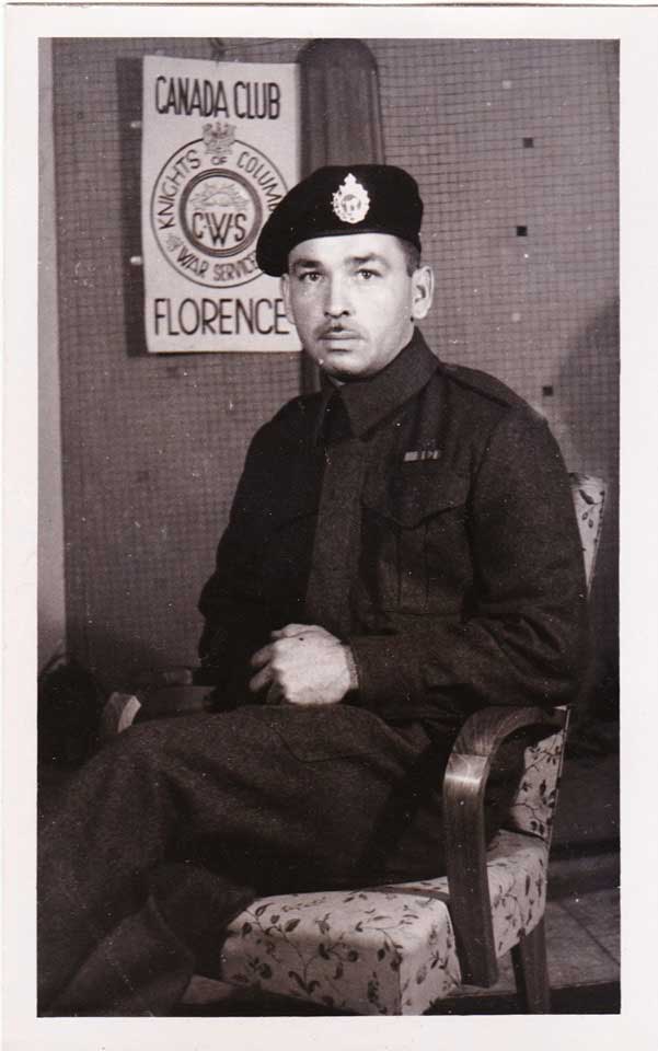  A Black and white photo of a man wearing an army uniform and beret. Behind him, a sign that reads Canada Club. Knights of Columbus War Service. Florence.