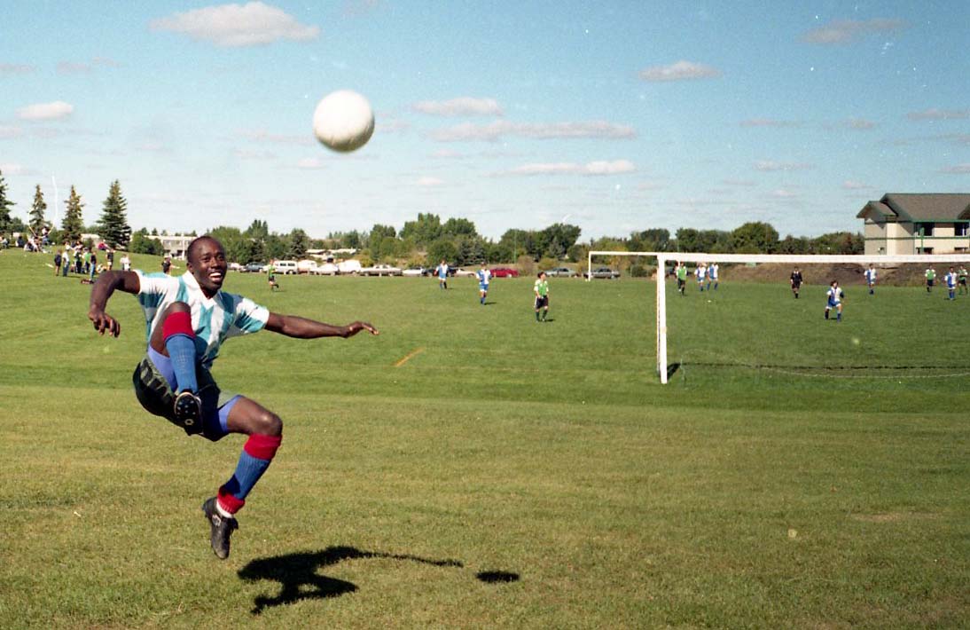 A black man smiles mid-air after kicking a ball, in bright sun on a large field with an active soccer pitch behind him.