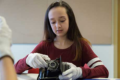 A young girl looks at a camera she is holding with gloved hands.