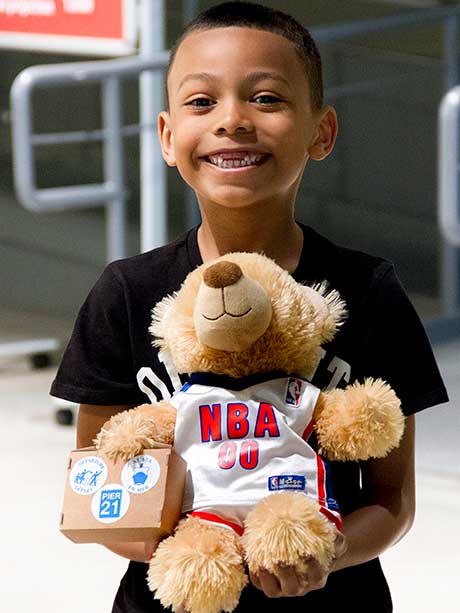 A smiling young boy is holding a teddy bear dressed as a basketball player.