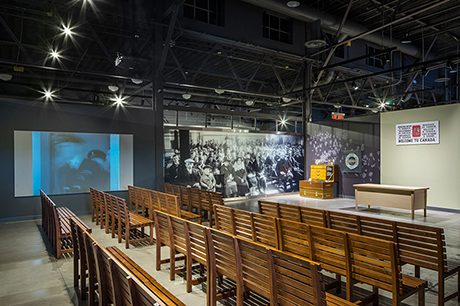 Big hall with wooden benches and large pictures on the wall and a screen showing Pier 21 images.
