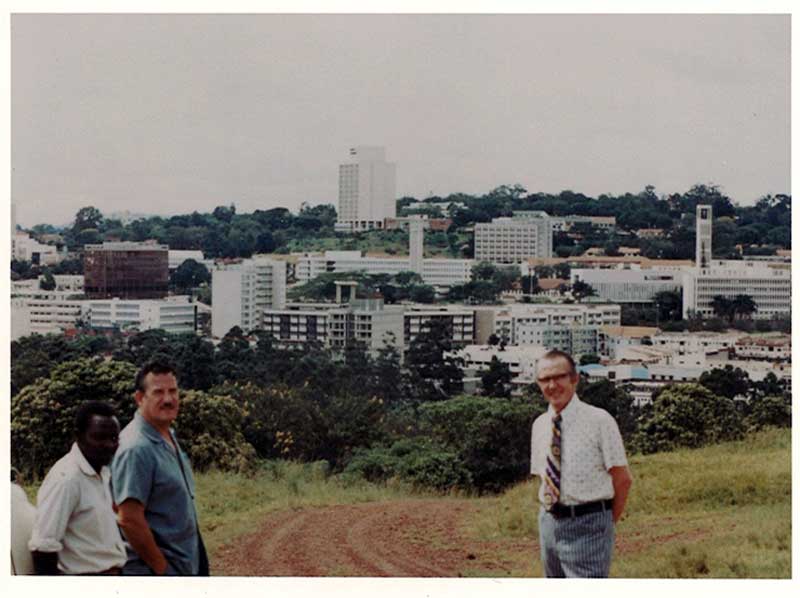 Three men stand on a dirt road with a small city in the background.
