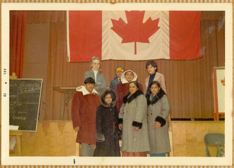 A family wearing warm winter clothing stand in front of a large Canadian flag hanging on the wall.