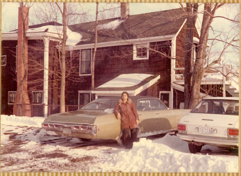 A young woman leans against a car in her driveway, there is a lot of snow on the ground.