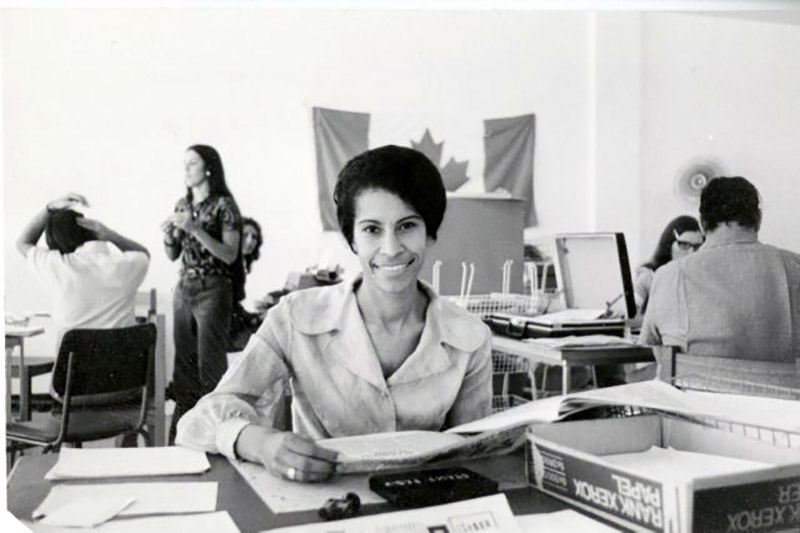Young woman seated at a desk is smiling and looking at documents. The Canadian flag hangs on the wall behind her.