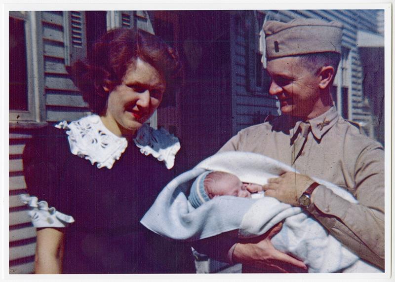 A young woman in blue stands next to a uniformed man who cuddles a baby in his arms.