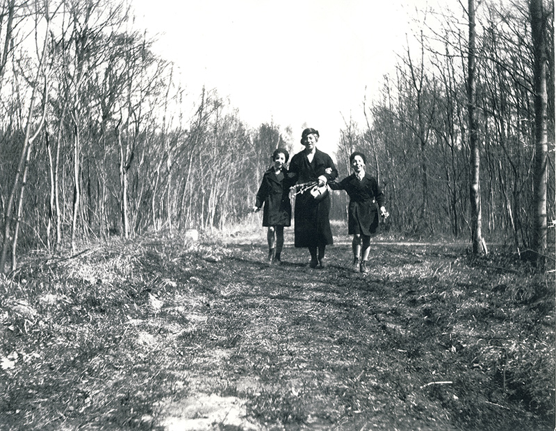 A woman and two children happily walk along a grassy path in the woods.