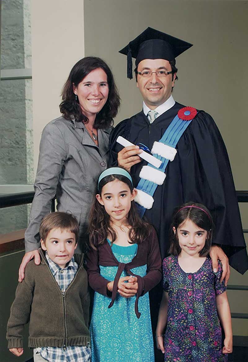A young man in graduation cap and clothing holds his degree proudly as he is surrounded by his wife and three children.