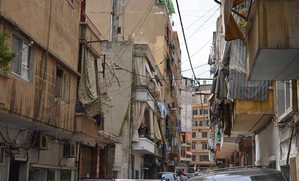 A cramped city street with cars parked on either side and all the homes have laundry drying on the clothelines.