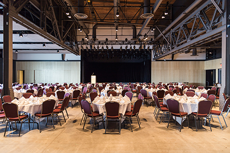 A big hall with lots of round dining tables covered with white tablecloths and there are six red chairs around every table.