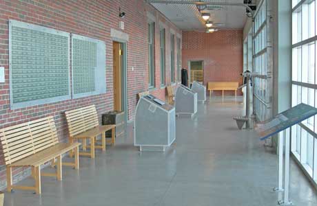 A room with a red brick wall on one side and floor to ceiling windows on the other, there are benches for sitting and green plaques on the wall.