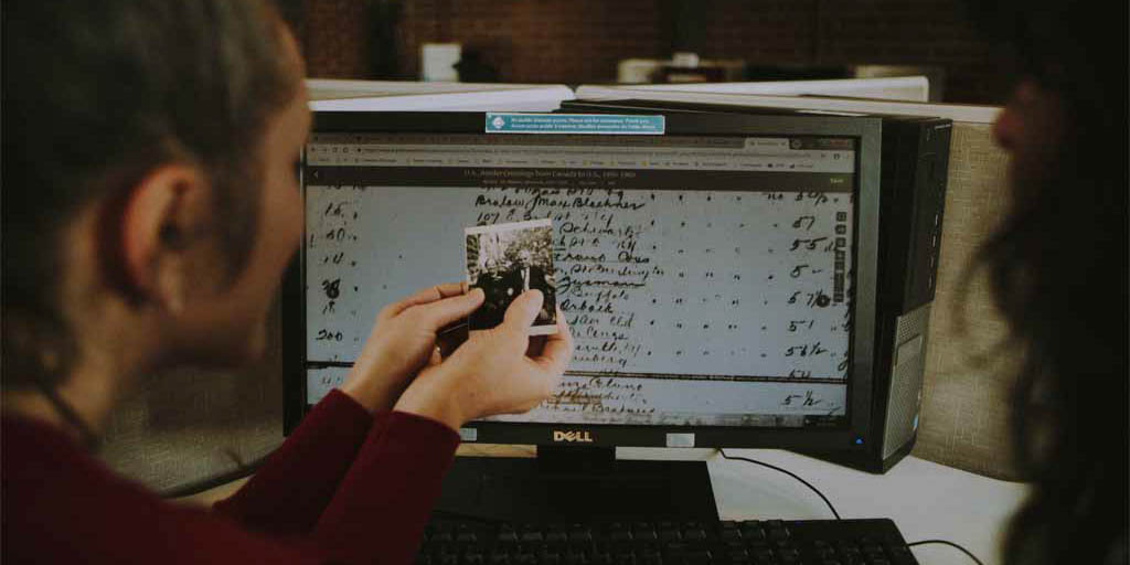 A woman holds up a black and white photo while looking at a screen showing an old handwritten document.