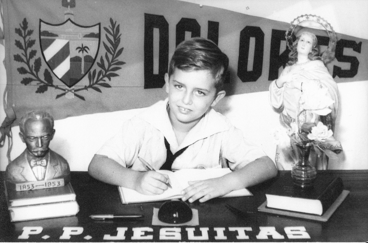 A young Rafael Alcolado, aged 6 or 7 sits at a desk doing schoolwork. A banner on the wall behind him says Dolores.