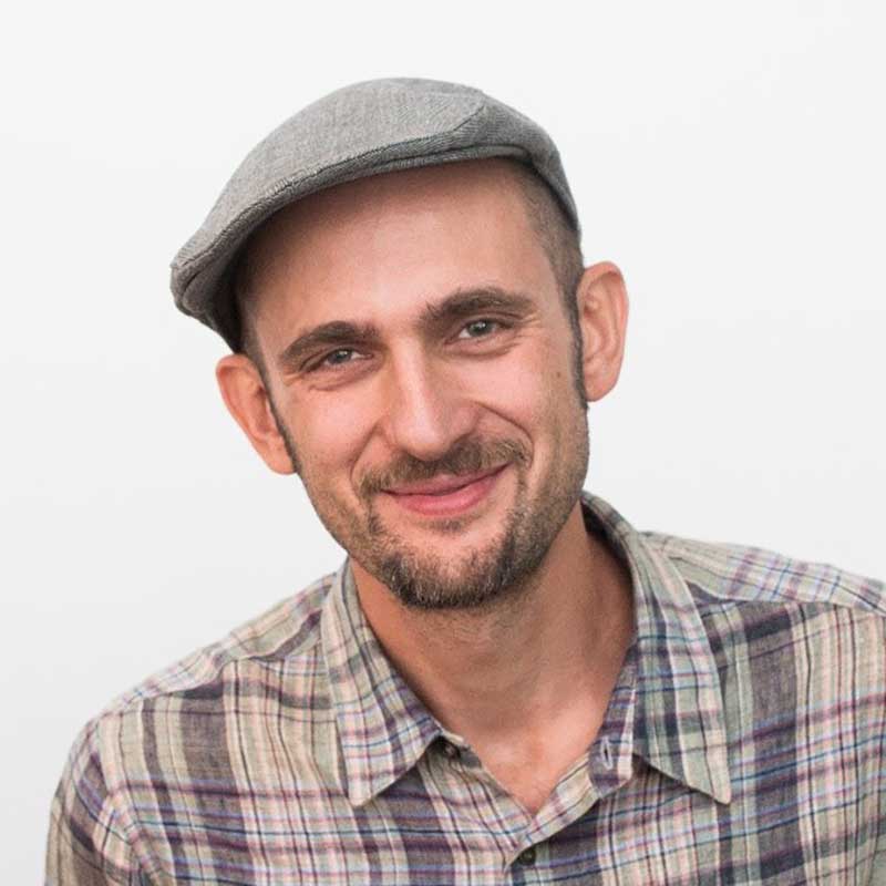 A man in a flat cap with a plaid shirt and a close-cropped beard smiles at the camera.