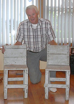 An older man kneels, smiling, behind two wooden child-sized chairs.