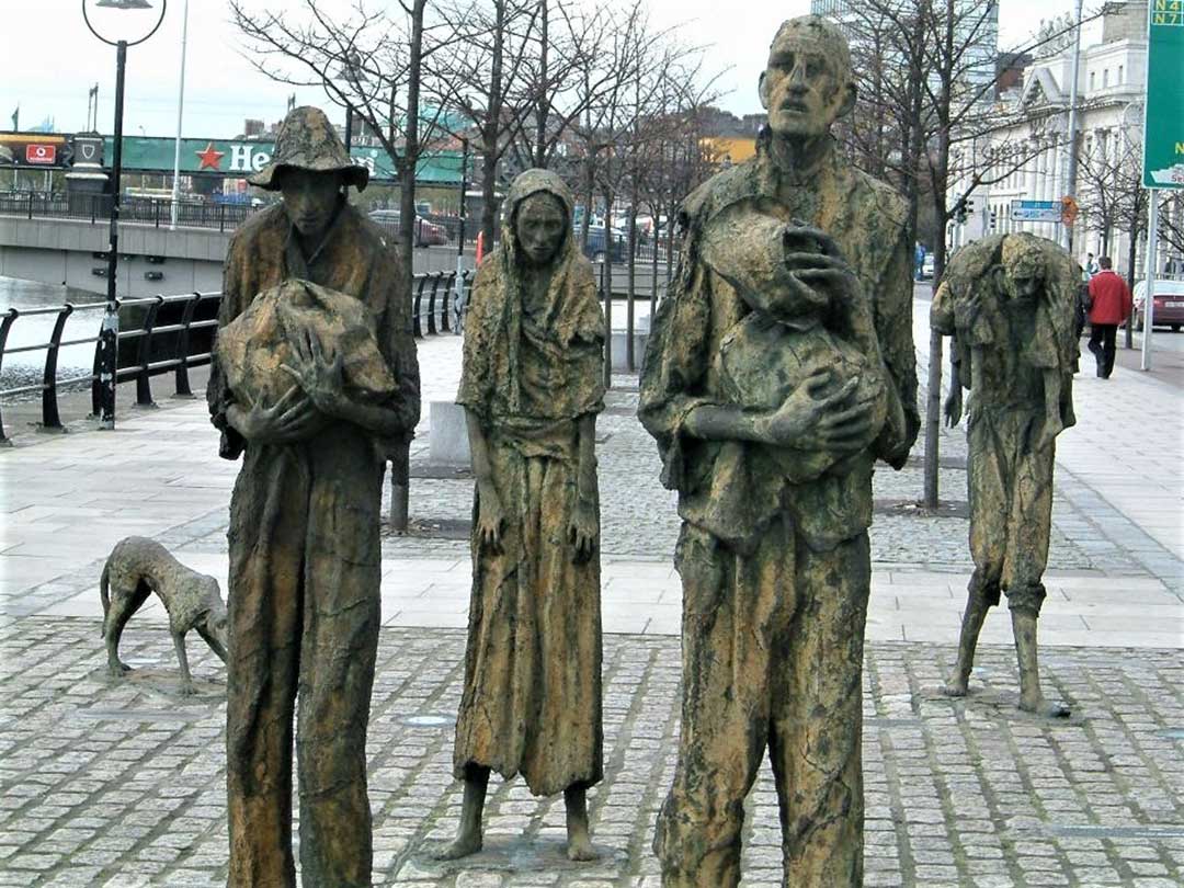 Bronze-cast statues of emaciated walkers on a wide city sidewalk, one figure carries a body over his shoulders.