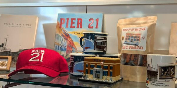 A glass shelf with various Pier 21 merchandise on it, including a hat, play bricks, mugs, and books.