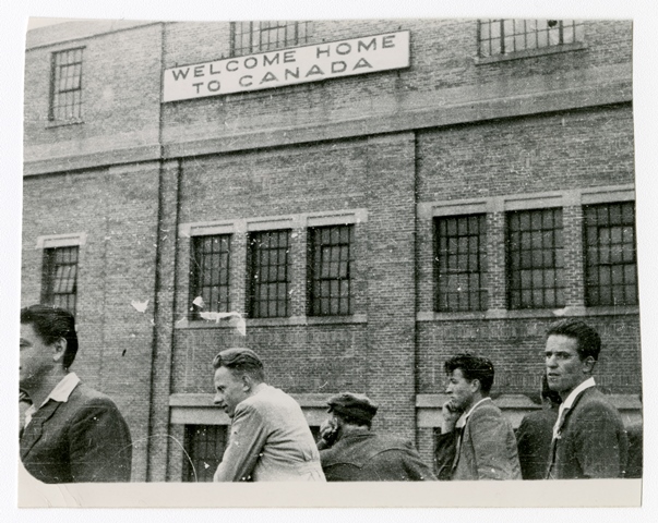 Black and white photo of young white men in front of a sign on the exterior wall of Pier 21 that reads Welcome Home to Canada.