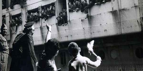 Old black and white war-time photo, showing a troop ship docking with hundreds of waving soldiers and a little boy and his mother waving back at them from the platform, while a serviceman looks on.