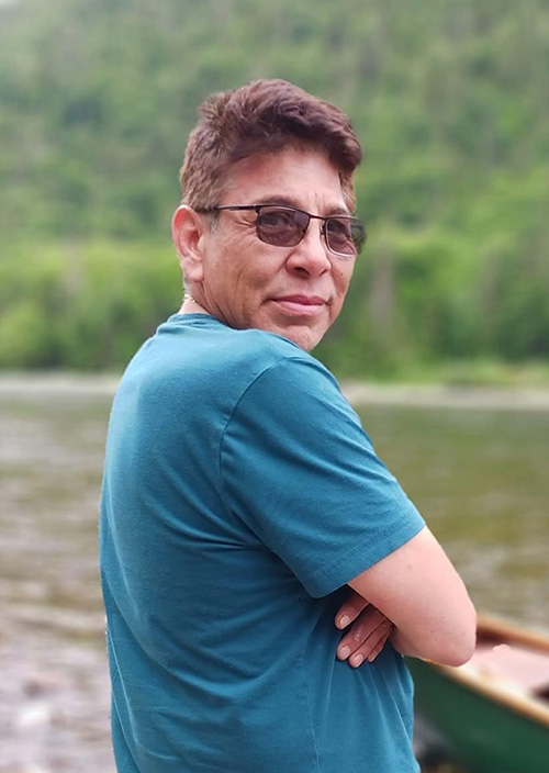 A man stands at a river by a canoe looking back towards the camera over his shoulder.