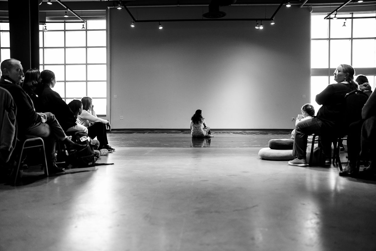 A woman sits on the floor, facing away from an audience.