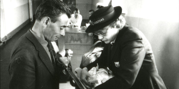 In an old black and white photo, a customs official rummages through the bag of a man in a suit.