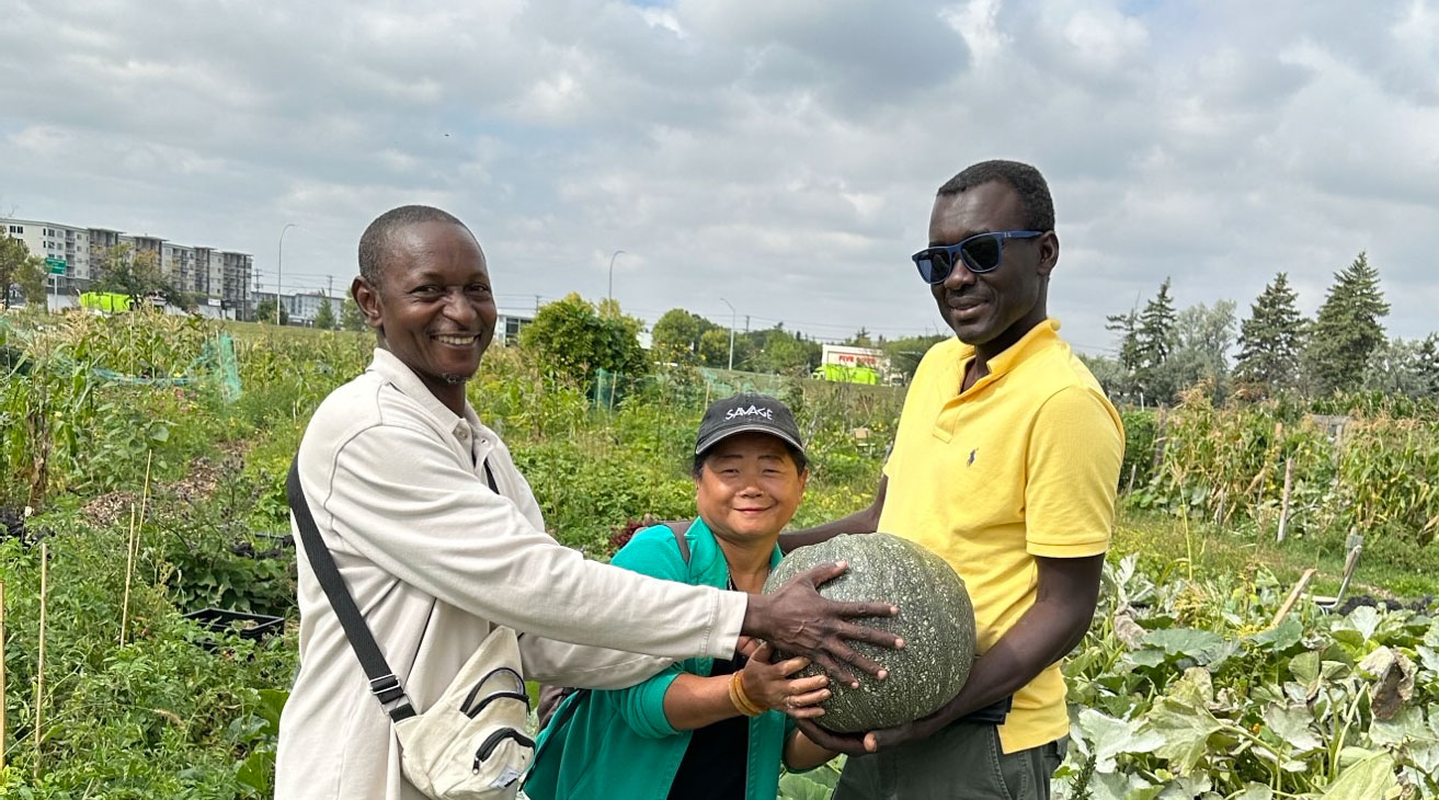 Two tall African men and a short Asian woman hold a green gourd in a large vegetable garden with apartment buildings in the background.