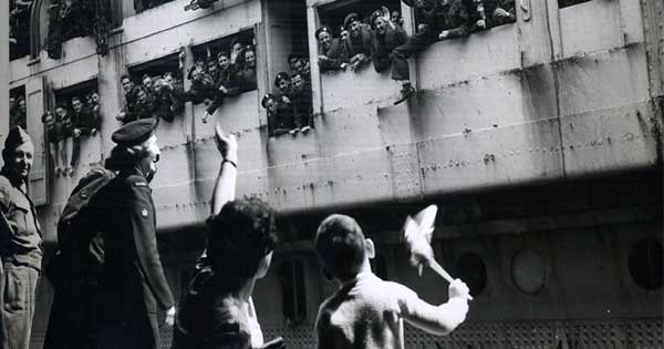 Old black and white war-time photo, showing a troop ship docking with hundreds of waving soldiers and a little boy and his mother waving back at them from the platform, while a serviceman looks on.