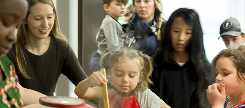 A young girl stirs a steaming pot with a wooden spoon in a room full of people.