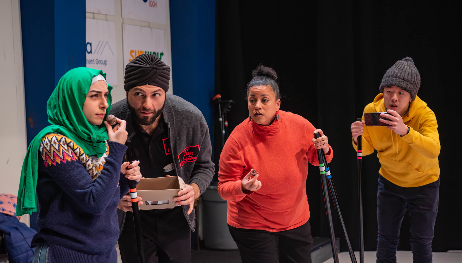 Three curlers, including a South Asian man in a turban, a Black woman, and an East Asian man, attentively watch a young Middle Eastern woman in a hijab eat a Timbit.