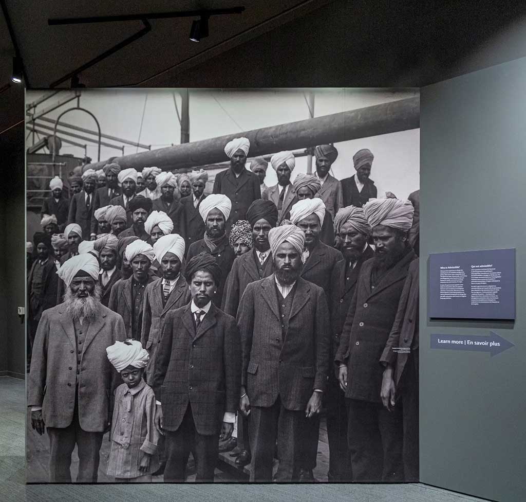 On a wall in the Museum, a large print of a black and white photo of around 30 men and one small boy in turbans aboard a ship. They look at the camera.