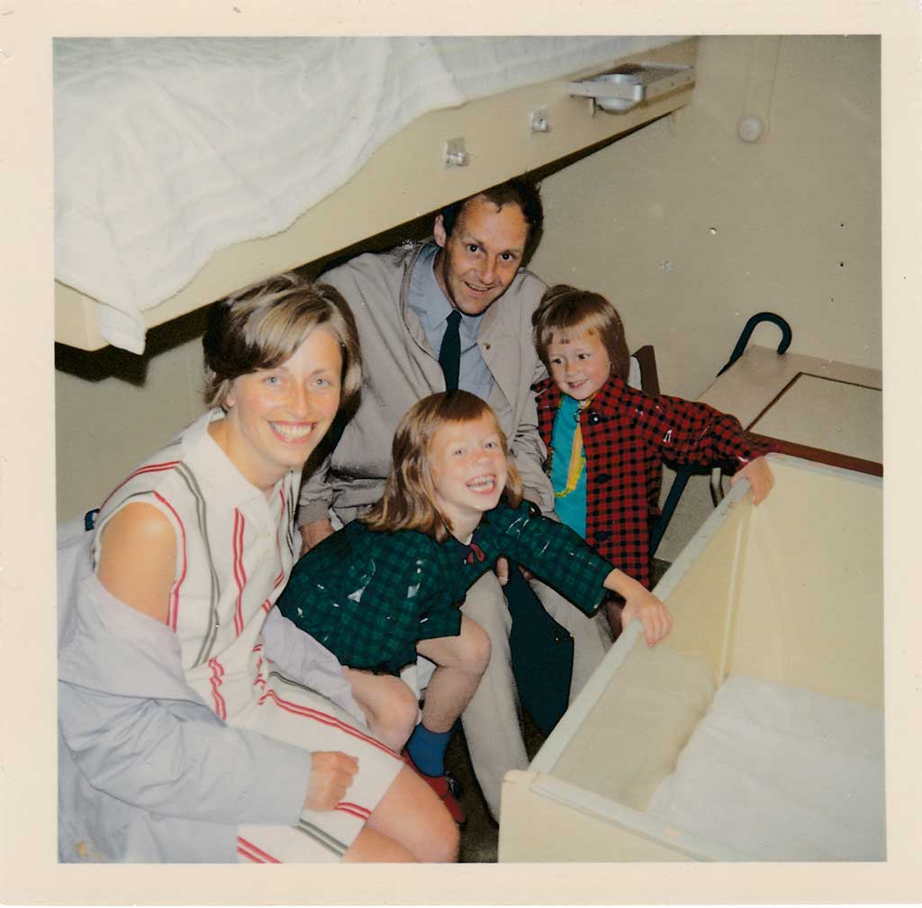 A father, mother, and two girls under 10, dressed in 1960's clothing, sit on the bottom bunk in a crowded ship cabin. It's tight but they are smiling.