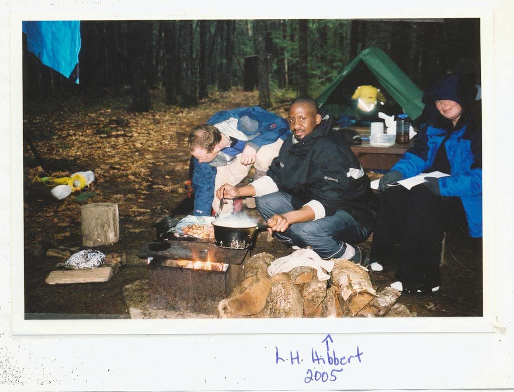 Color photo of Lyndon Horrace Hibbert camping in 2005. He is squatting next to a fire, cooking and wearing rain gear. Two people sit beside him and there is a person and tent in the background.