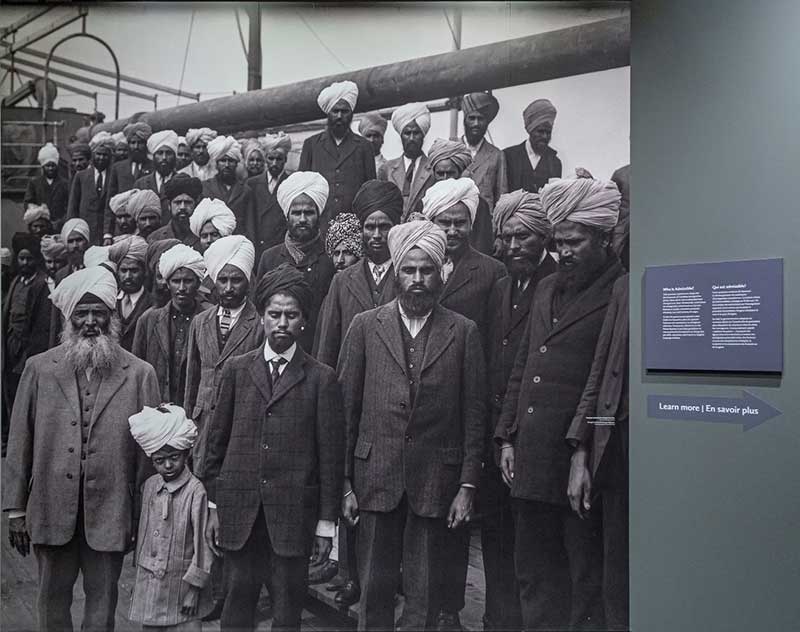 On a wall in the Museum, a large print of a black and white photo of around 30 men and one small boy in turbans aboard a ship. They look at the camera.