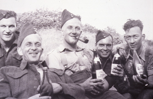 5 men in army uniforms, some holding bottles of beer, smile for the camera. Cliff is centre with a pipe in his mouth.