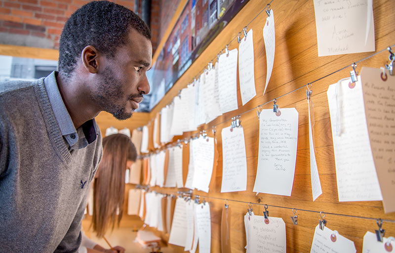 A black man wearing a collared shirt and sweater reading handwritten notes on paper luggage tags hanging on a wall.