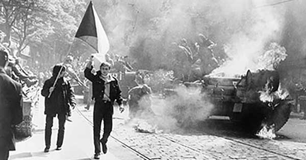 A black and white photo of two men on a sidewalk, one holding a Czechoslovakian flag, as a tank burns on the street beside them.