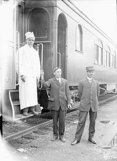 Three men, one in a chef's uniform, one in a suit and one in a train porter’s uniform stand outside a train car. 