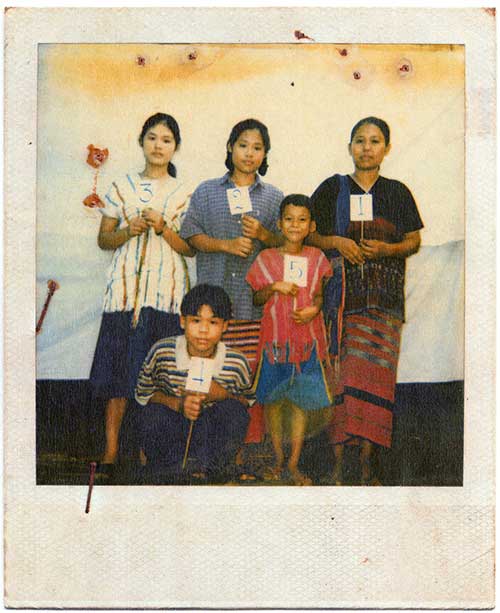 A polaroid photo of a family of 5, mother, two daughters and two sons, stand against a white tarp  backdrop each holding a small sign with a number, 1 to 5.