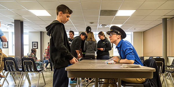 A boy stands at a table. On the other side of the table, a Museum staff member wearing the hat of a customs official listens attentively as the boy talks.