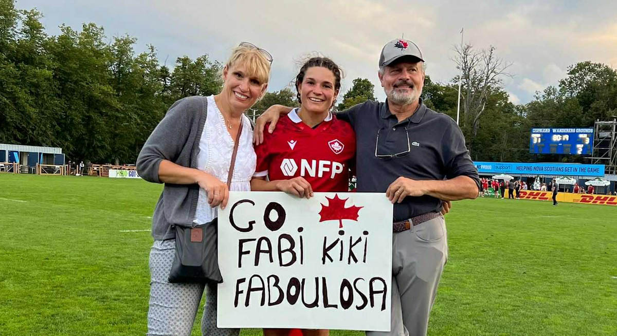 A middle-aged couple stands with a young woman wearing a Rugby Canada Uniform, on a field. They hold a sign that says Go Fabi Kiki Fabulosa.