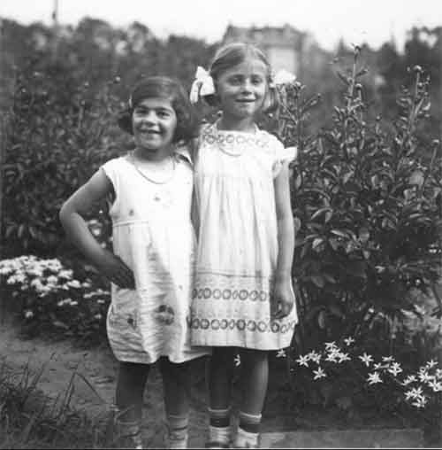 Two young girls in white dresses stand in a garden.