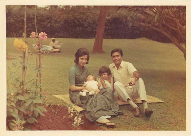  A family of four, including a newborn baby and girl of about 4, sit on a blanket on a well-trimmed lawn with flowers in the foreground.