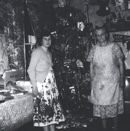 Black and white photograph of two women, one older wearing an apron, standing next to a decorated Christmas tree