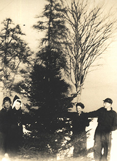 A black and white photograph of two men, and woman and a boy, outside wearing coats and hats, laughing next to a very tall evergreen tree