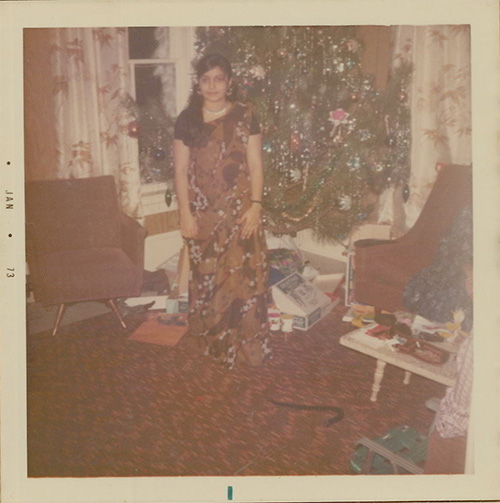 A faded colour photograph of a woman dressed in a sari standing in front of a decorated Christmas tree, opened boxes and packages surround the base of the tree