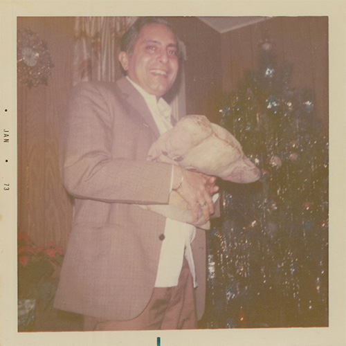 A faded colour photograph of a well-dressed man smiling next to a decorated Christmas tree, holding a full burlap sack of unknown contents