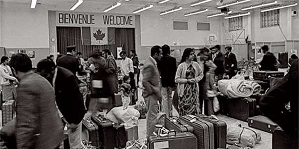 A large room crowded with People and suitcases. A Canadian flag and the word Welcome, in English and French appear on the back wall.