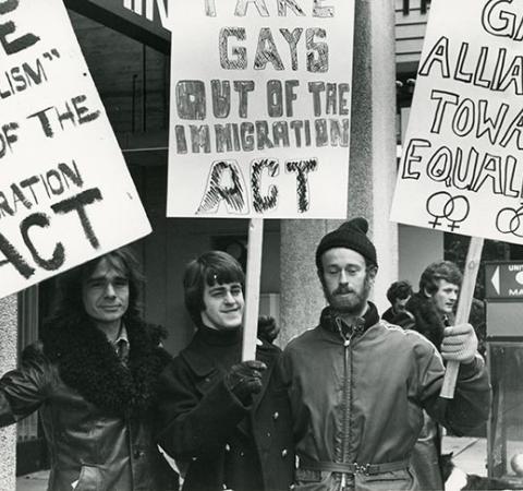 Three men with signs: Take homosexualism out of the Immigration Act, Take gays out of the Immigration Act, and Gay Alliance Towards Equality. They are protesting in front of a commercial building and are dressed for cool weather." 