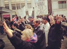 A crowd of people walk through a city street holding protest signs, one sign reads I wanna stay in Canada.